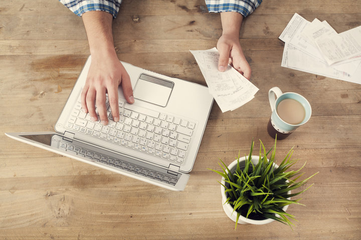 Hands holding a bill while typing on open laptop sitting on a wooden surface. Next to the laptop sits a mug full of coffee and a small green plant in a white flower pot. 