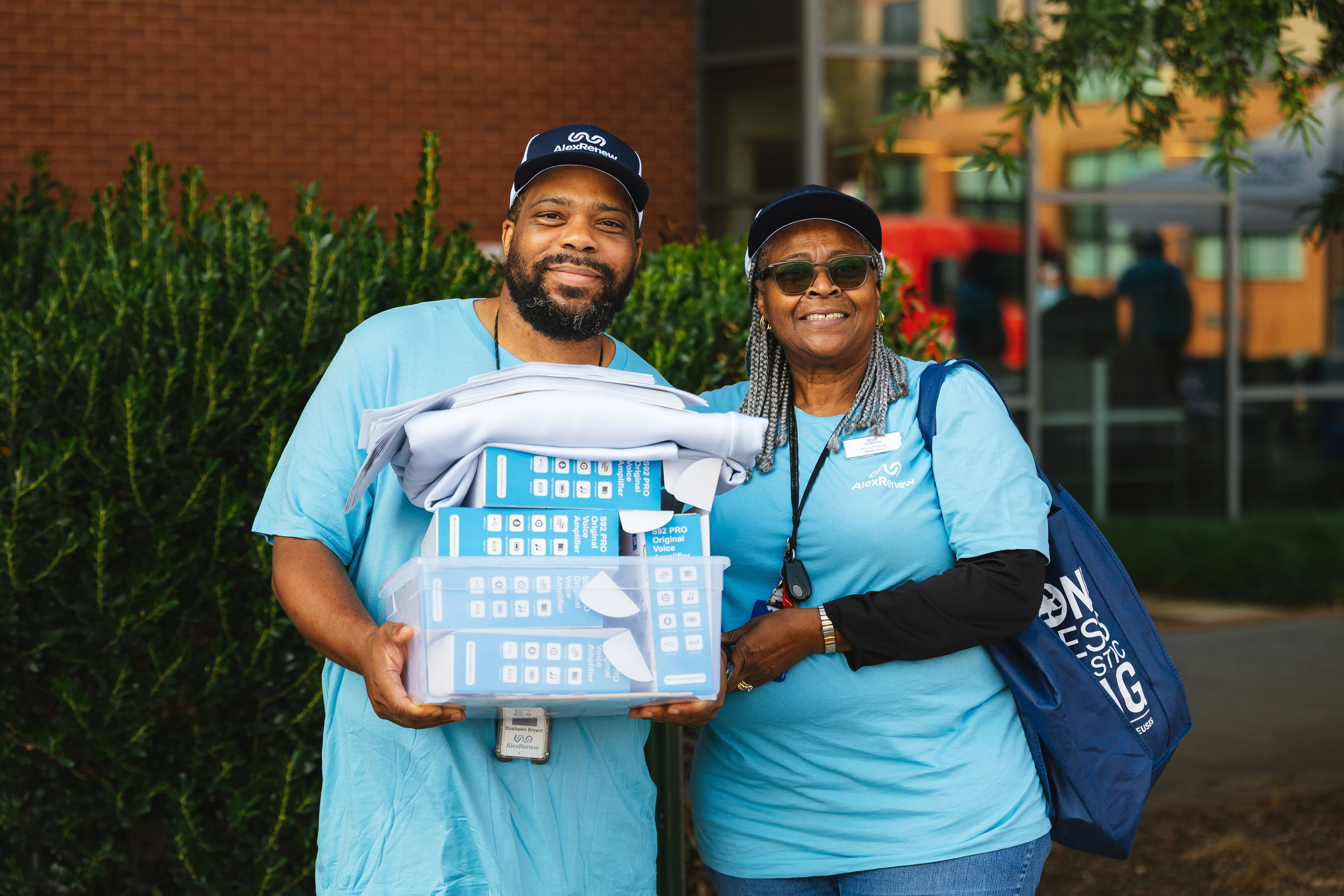 A Black man and woman in blue AlexRenew t-shirts smile while holding a box of equipment for the Open House.
