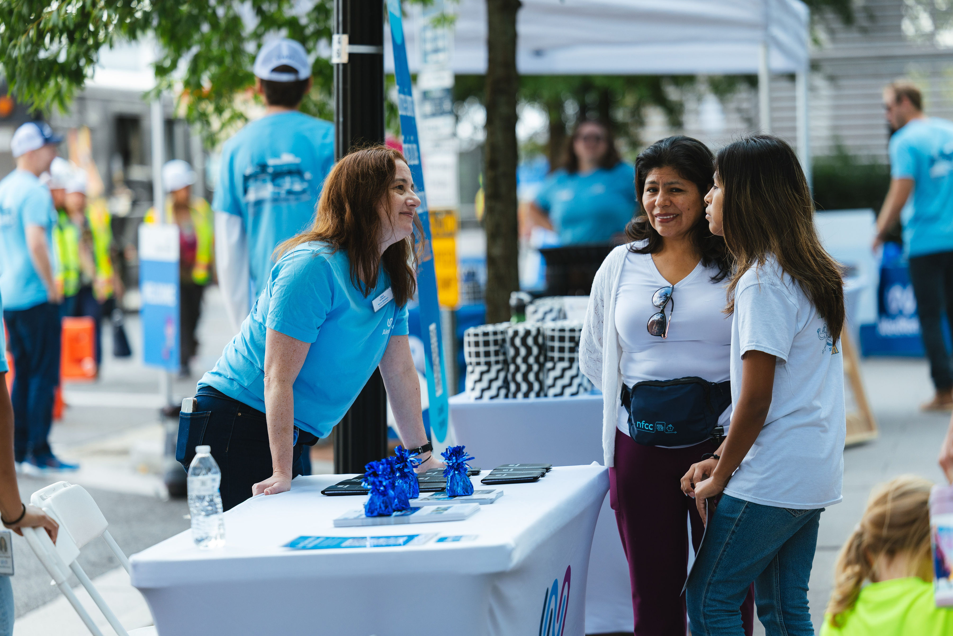 A woman in a blue AlexRenew t-shirt leans across a table with promotional materials to talk to two women. In the background more people mill around the Open House.