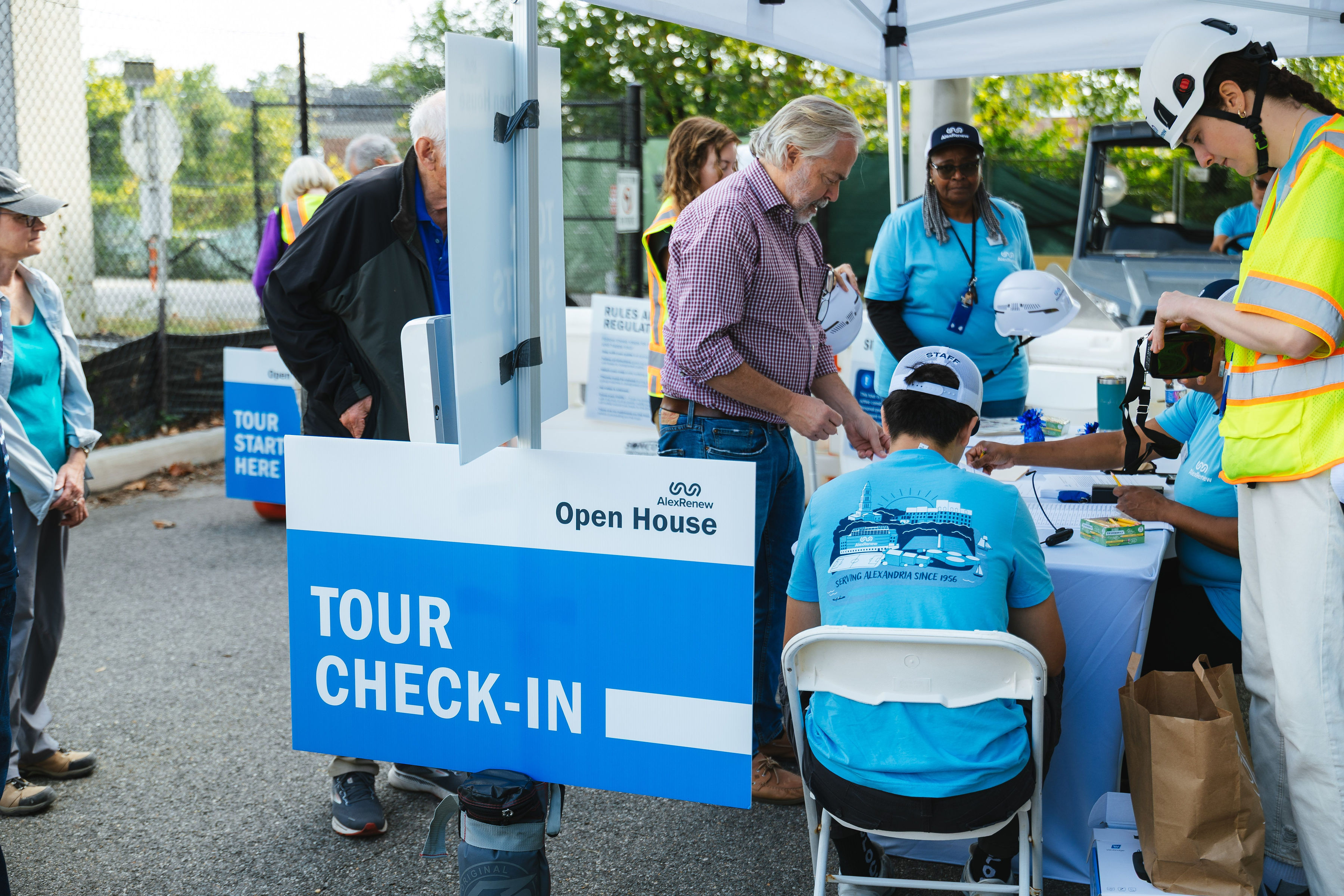 A blue "Tour Check-In" sign in the foreground sits in front of a table where an elderly man in a checkered shirt talks to AlexRenew staff in safety vests.