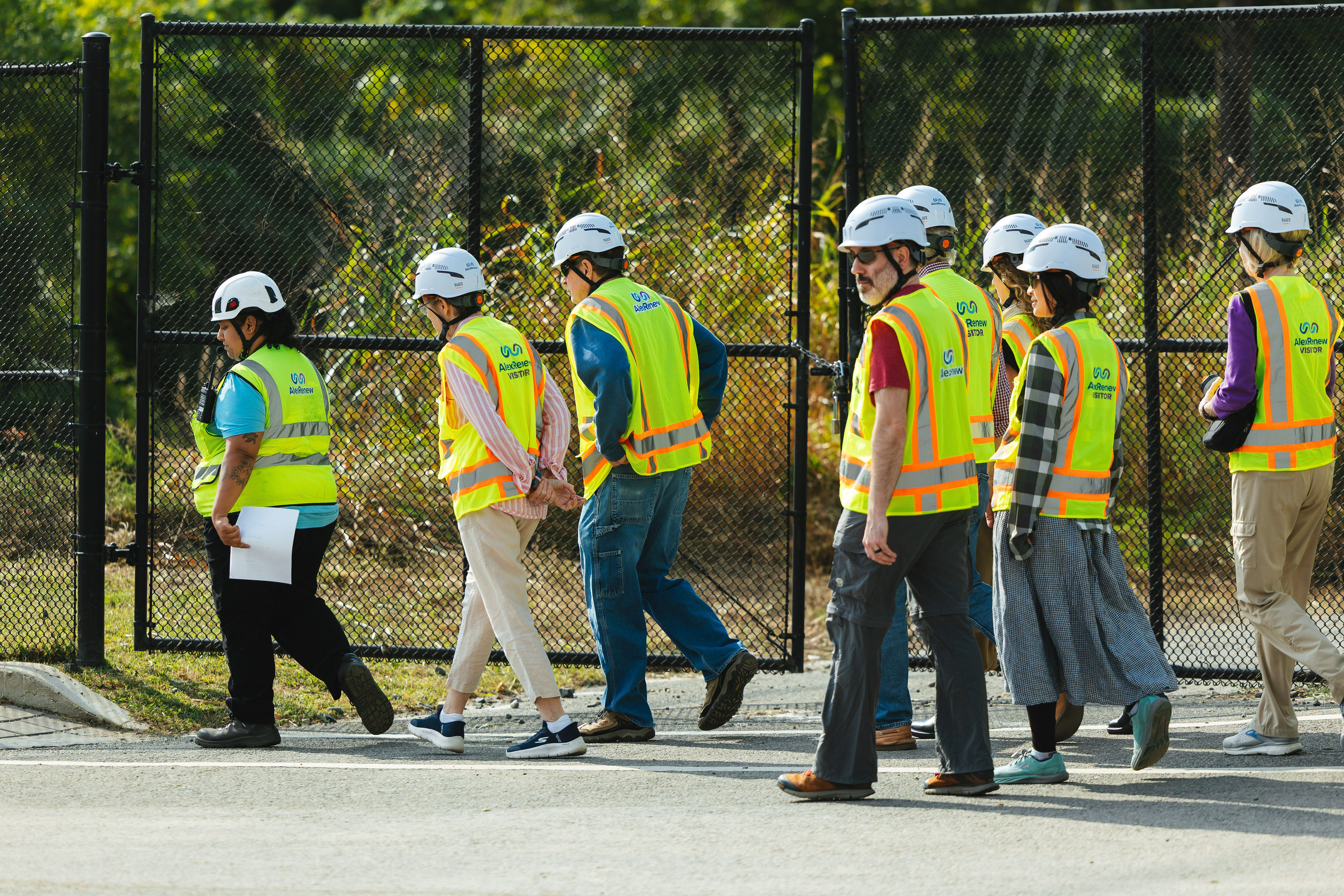 Several adults in hard hats and safety vests walk in front of a fence and trees