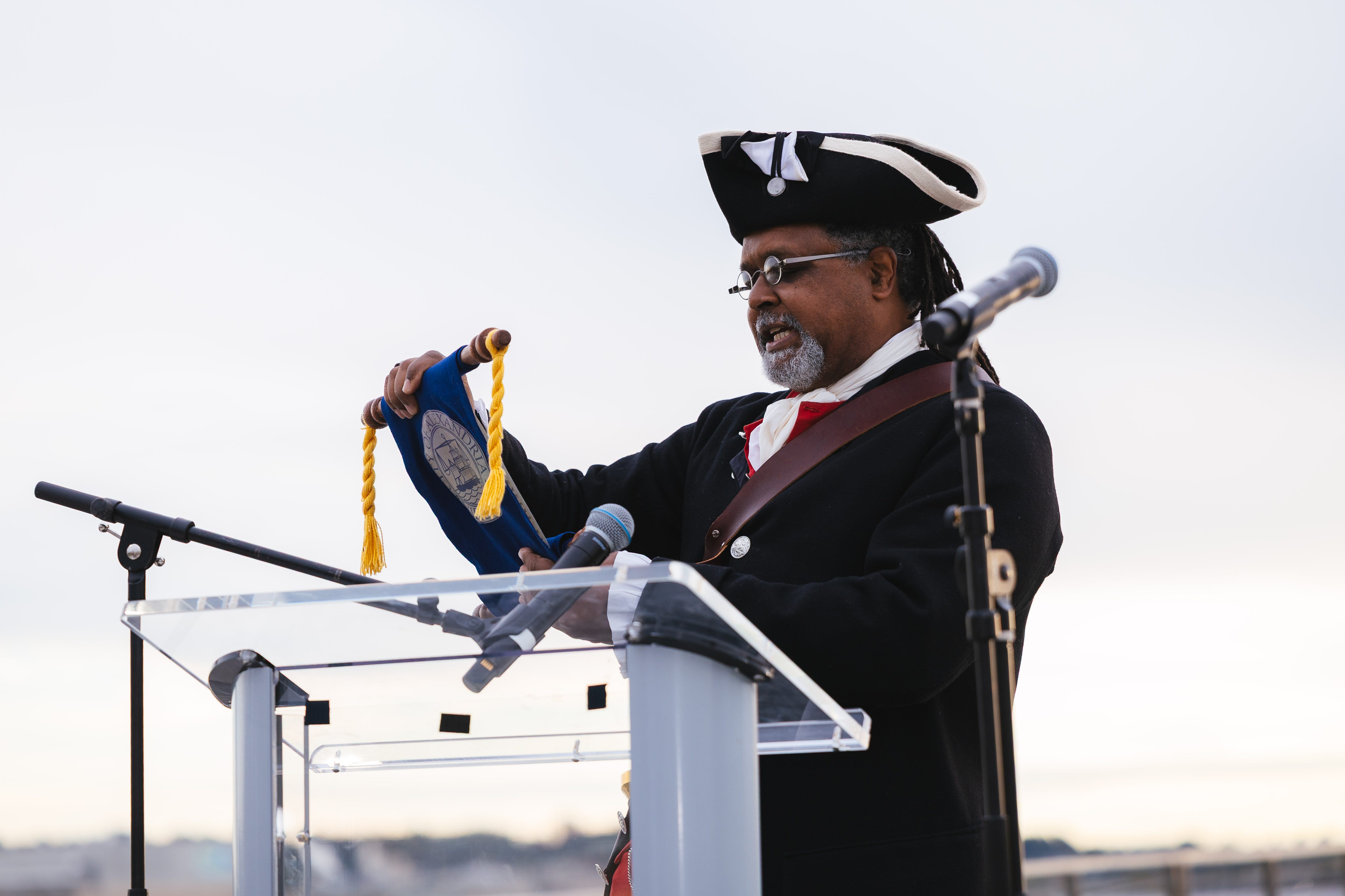 A man dressed in a revolutionary war costume is the town crier. He reads a proclamation from a scroll behind a podium.