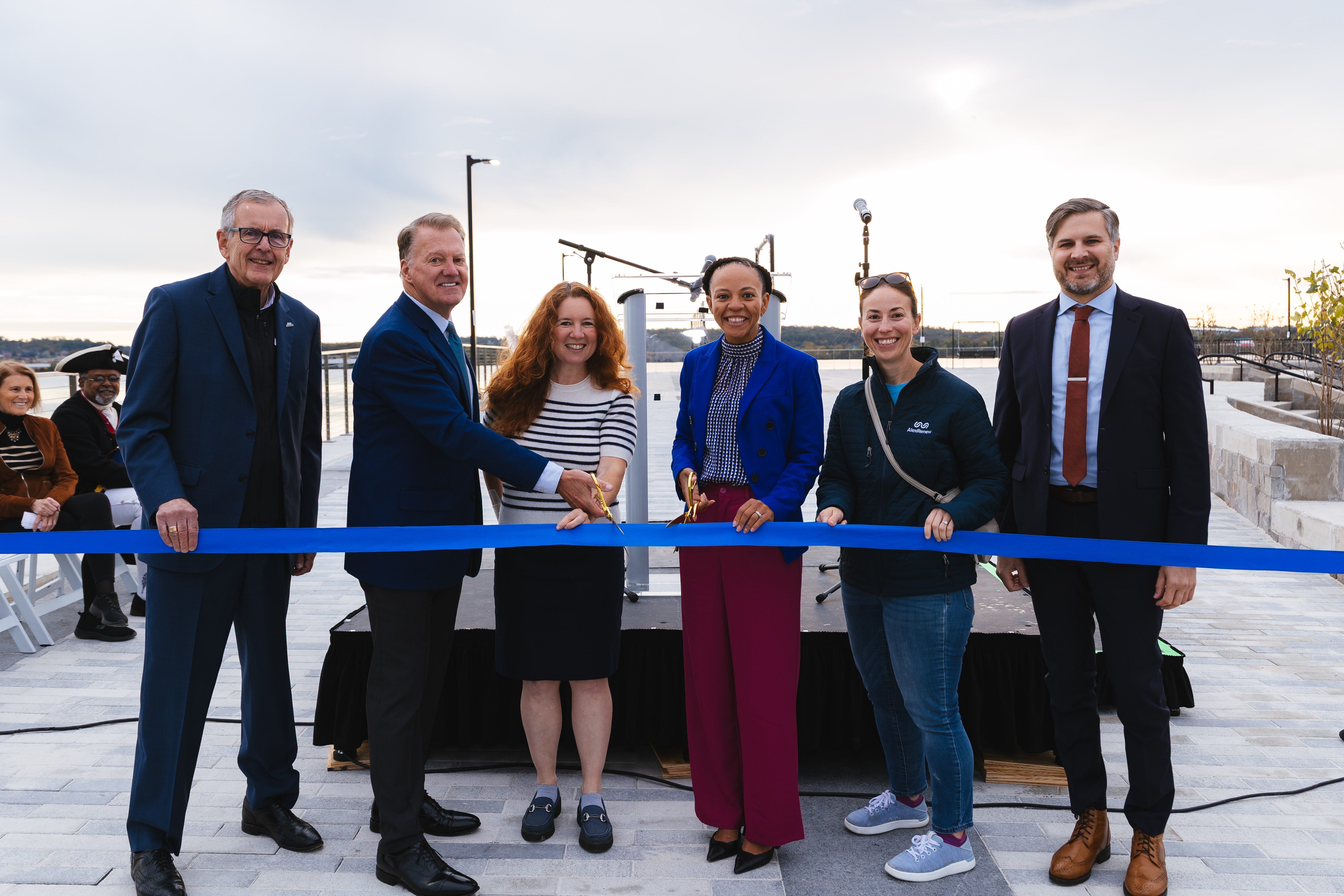 Six men and women in business attire stand behind a blue ribbon. The woman in the center, the mayor, holds a pair of scissors ready to cut the ribbon.