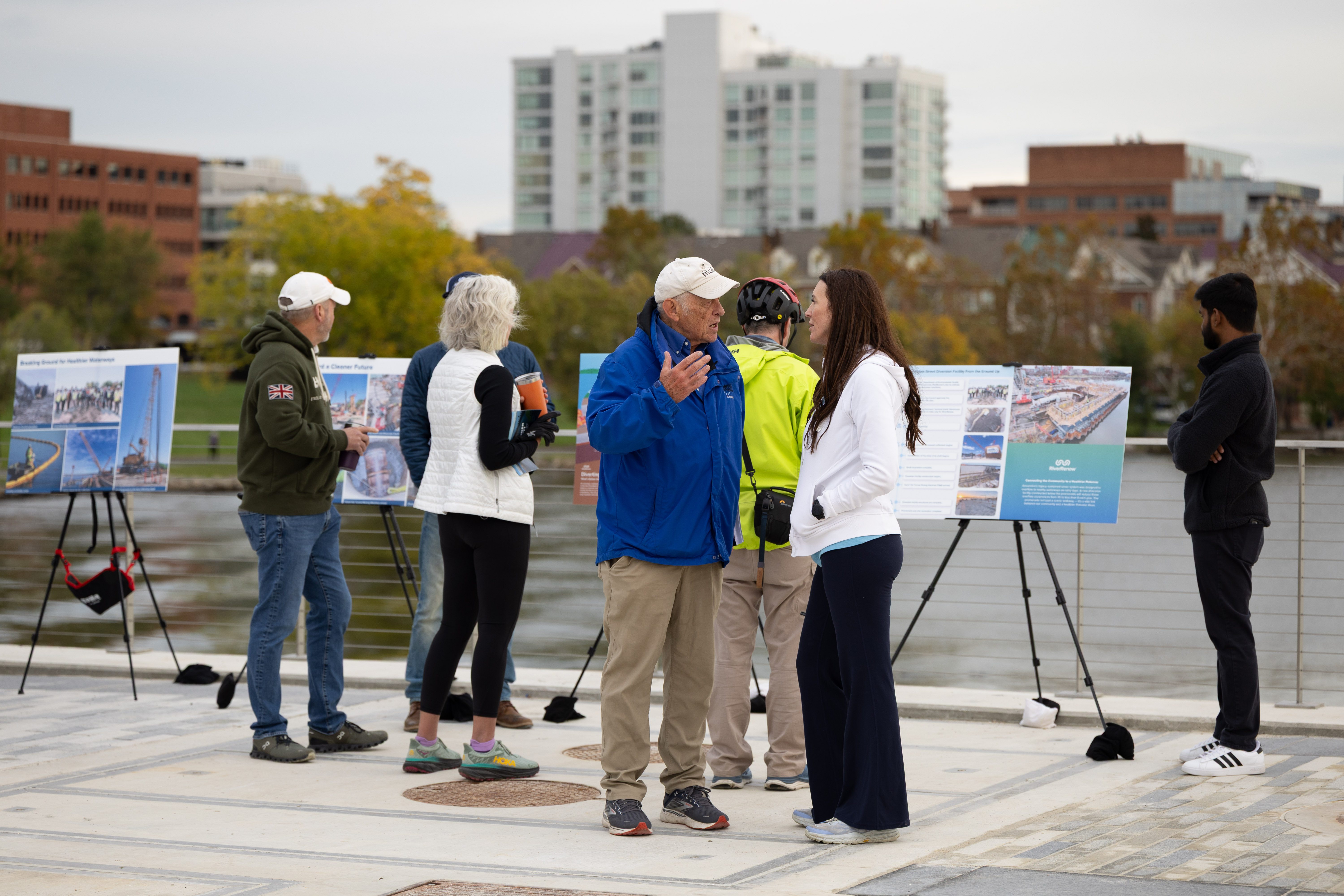 People in light jackets talk in front of informational signs placed along the edge of the promenade, with the river in the background.