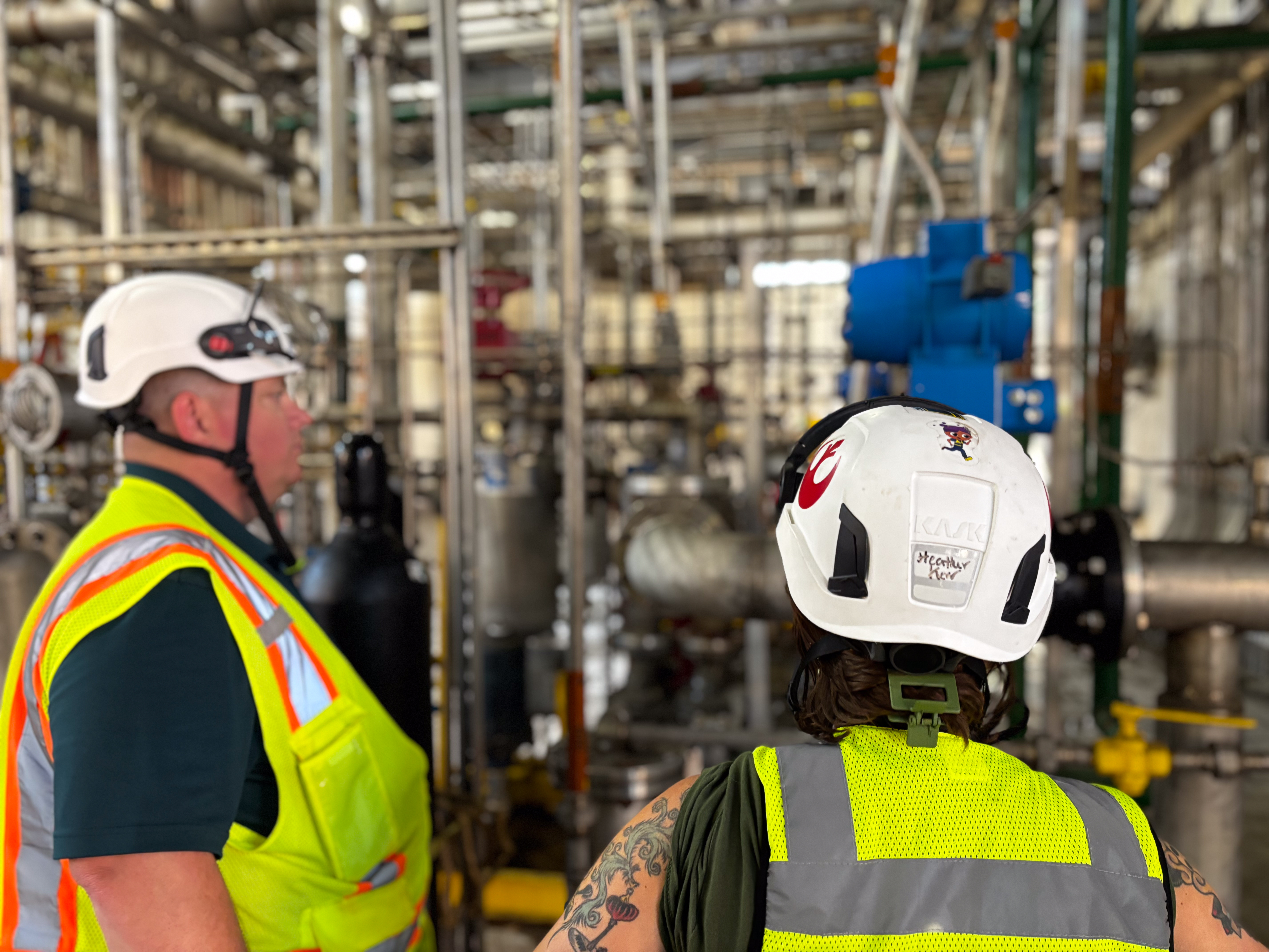 Two employees in hard hats and safety vests inspect a pipe.