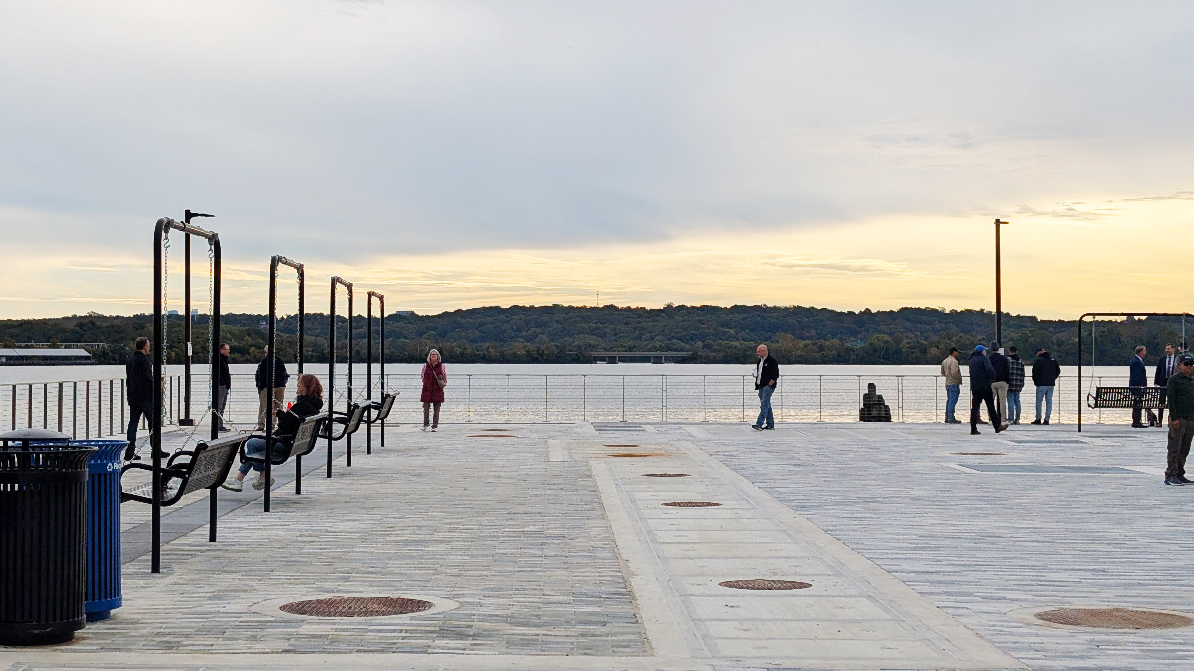 The paved promenade at sunrise overlooking the Potomac River. A few people stroll around the edge of the promenade or sit on benches overlooking the water.