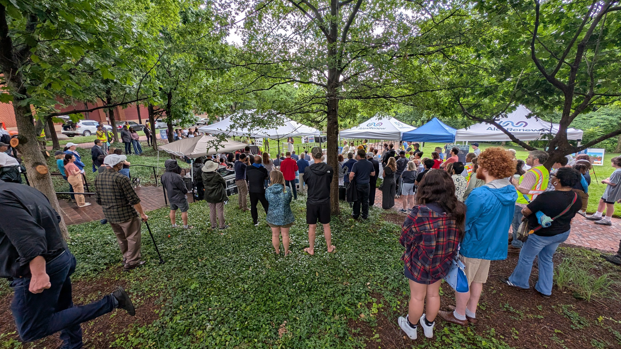 People gather around popup tents in a park, listening to a speaker.