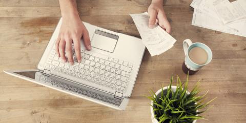 Hands holding a bill while typing on open laptop sitting on a wooden surface. Next to the laptop sits a mug full of coffee and a small green plant in a white flower pot. 