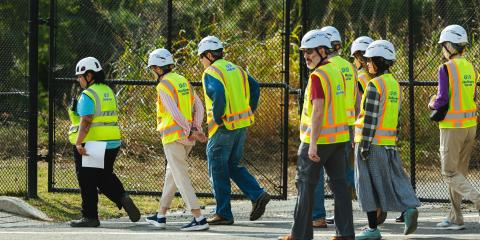 Several adults in hard hats and safety vests walk in front of a fence and trees