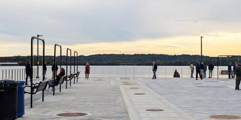 The paved promenade at sunrise overlooking the Potomac River. A few people stroll around the edge of the promenade or sit on benches overlooking the water.